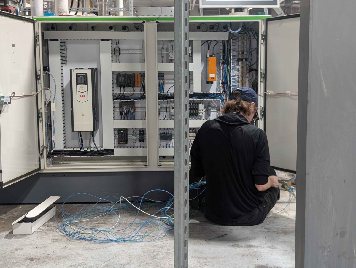 Technician sitting on the floor pulling control wire into an open panel fitted with an ABB drive, PLC I/O and terminal blocks