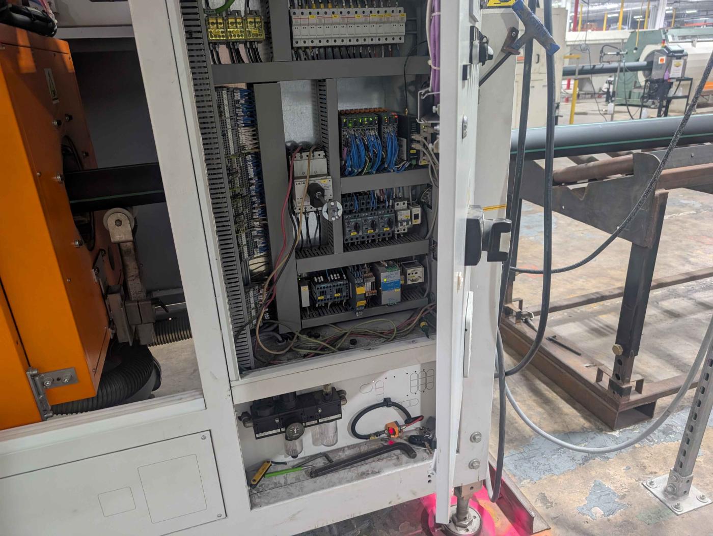 Open machine control cabinet on a plant floor showing circuit breakers, terminal blocks, networked I/O modules, main disconnect, motor protection breakers and a safety relay